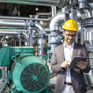 Power plant engineer standing by gas engines inside energy production factory and checking results on digital tablet.